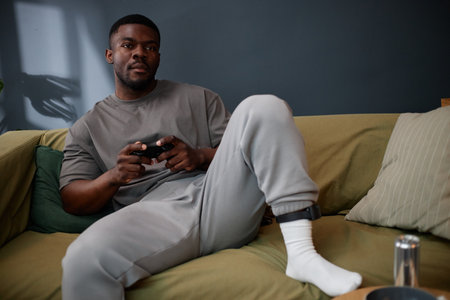 Young adult Black man sitting on sofa wearing electronic ankle monitor, holding game controller looking away from camera representing concept of house arrest and electronic monitoringの写真素材
