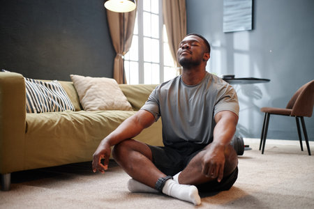 Young adult Black man sitting cross legged on carpet meditating with eyes closed wearing electronic ankle monitor in living room during house arrest, sunlight streaming through windowの写真素材