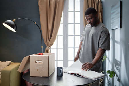 Young adult Black man standing near table turning blank pages of photo album, with cardboard box and coffee mug nearby in home setting during house arrest situationの写真素材