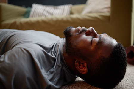 Young adult Black man lying on floor with eyes closed, appearing calm and relaxed in home setting,...