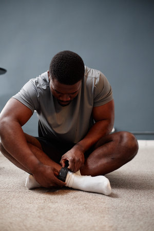 Black young adult man sitting cross legged on carpeted floor attaching electronic ankle monitor to socked ankle suggesting house arrest or electronic monitoring situationの写真素材