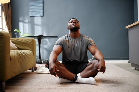 Black young adult man sitting cross legged on carpeted floor in living room meditating with eyes closed during house arrestの写真素材