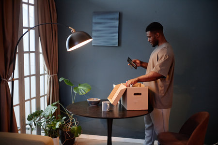 Young adult Black man standing near window using smartphone while unpacking cardboard box on round table in modern room, house arrest context implied by indoor settingの写真素材