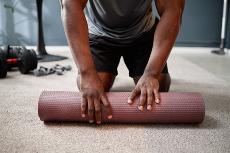 Black young adult man kneeling on carpeted floor rolling up yoga mat with dumbbells and resistance bands nearby, concept of exercising at home during house arrestの写真素材
