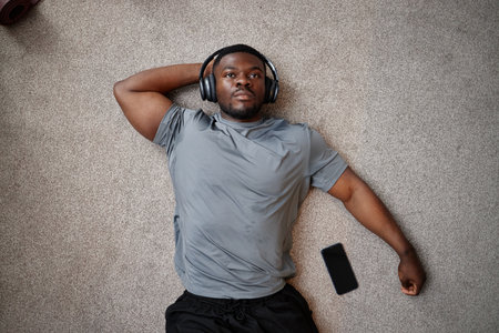 Young adult Black man lying on carpet listening to music with wireless headphones, relaxing with smartphone nearby, appearing thoughtful and calmの写真素材