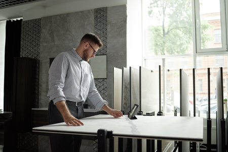 Caucasian man standing at large table examining architectural plans or samples in modern office, using digital tablet for reference, natural light coming through windowの写真素材