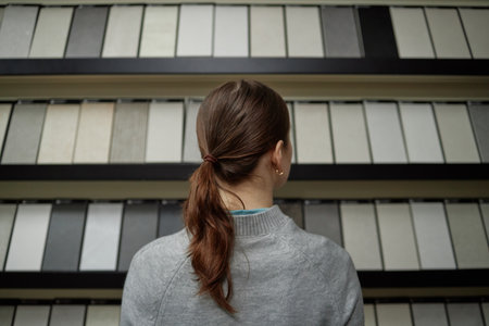 Caucasian young adult woman standing with back to camera facing display of rectangular samples arranged on shelves, brown hair tied in ponytail, wearing sweaterの写真素材
