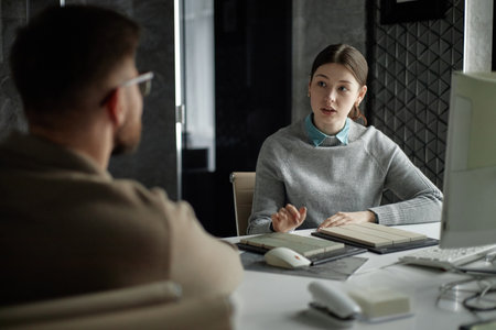 Young Caucasian woman discussing project details with bearded Caucasian man in modern office setting, both sitting at desk with computer and material samples visibleの写真素材