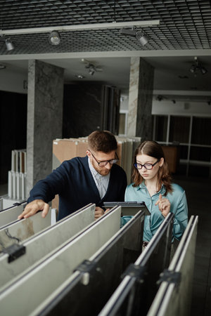 Caucasian man and woman examining large tile samples together in showroom, both wearing glasses and discussing options while using digital tabletの写真素材