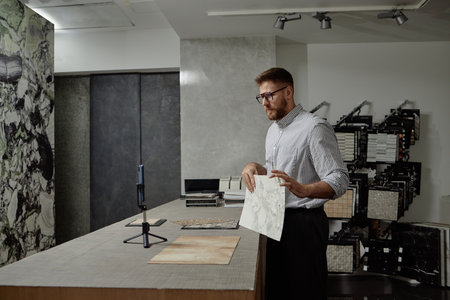 Caucasian man standing at showroom counter holding marble tile sample and gesturing while recording...