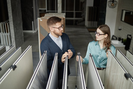 Caucasian young adult man and woman discussing and examining large tile samples in modern showroom, both standing between display racks and interacting with materialsの写真素材