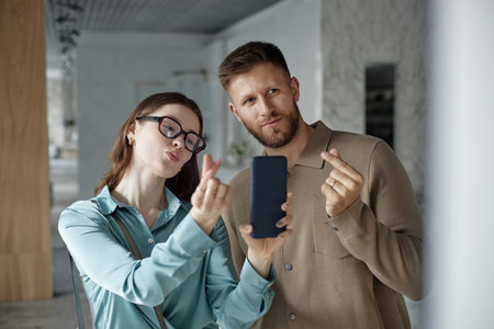 Caucasian young woman wearing glasses and Caucasian young man making finger heart gestures while taking selfie with smartphone, both looking at camera and smiling playfullyの写真素材