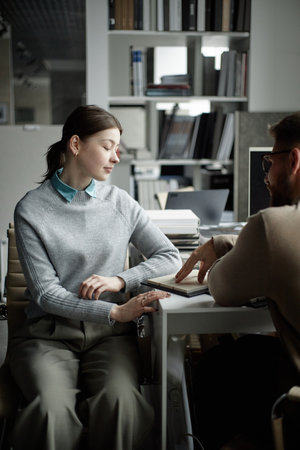 Caucasian young adult woman sitting at desk listening to Caucasian young adult man explaining information while pointing at notebook in modern office settingの写真素材