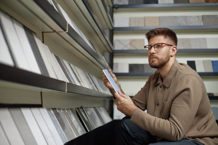 Caucasian man wearing glasses selecting ceramic tiles from display shelves, holding two samples in hands, examining surface texture and comparing options in storeの写真素材