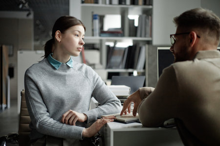 Caucasian young adult woman listening attentively to Caucasian young adult man during business conversation in modern office setting, both seated at desk with documents and computer visibleの写真素材