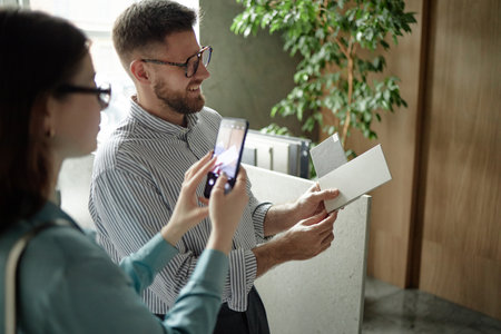 Caucasian young adult man smiling while holding samples, standing next to Caucasian young adult woman taking photo with smartphone in modern office settingの写真素材