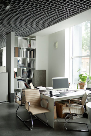 Modern office workspace featuring empty chairs surrounding desk with computer monitor, bookshelves filled with documents in background, large window letting in natural light, no people visibleの写真素材