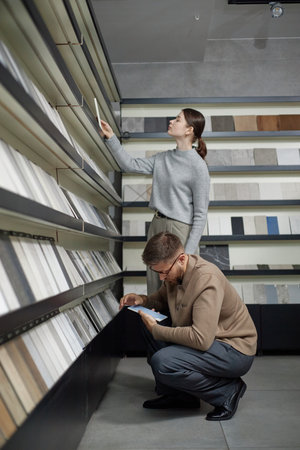 Caucasian young woman standing and selecting material sample from shelf while man crouching and examining catalog in modern showroom with various surface samplesの写真素材