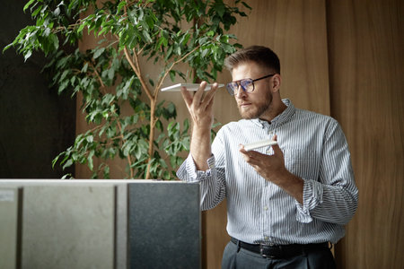 Caucasian young adult man wearing glasses holding two plates, standing indoors near large plant, examining plate closely with focused expression, office environment visibleの写真素材
