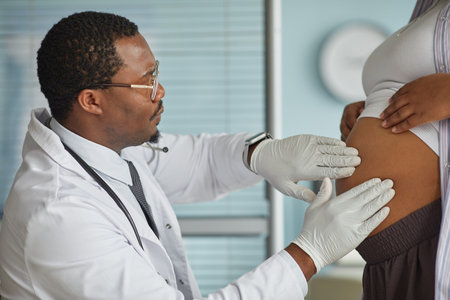 Black male adult doctor examining pregnant belly of Black woman in medical office, doctor wearing gloves and stethoscope, patient standing while doctor performing checkupの写真素材
