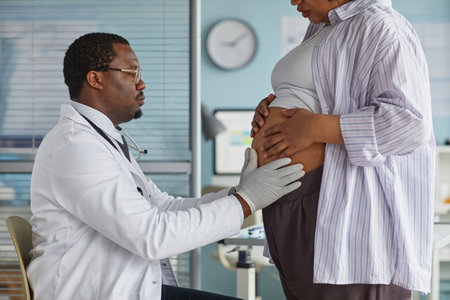 Black male doctor examining pregnant Black woman by palpating abdomen during prenatal checkup in medical office, both adults focused on healthcare assessment, clinical setting visibleの写真素材