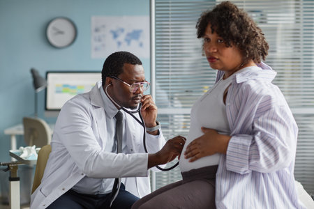 Black male doctor examining pregnant Black woman with stethoscope in medical office, middle aged physician focusing on prenatal care while expectant mother sitting on chairの写真素材