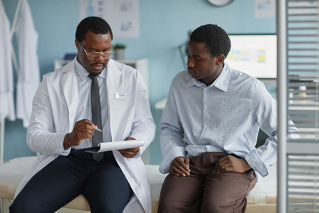 Black middle aged male doctor explaining medical information to young Black man patient during consultation in clinic office, both sitting on examination table and discussing healthの写真素材