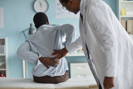 Black young adult man sitting on examination table holding lower back while Black male doctor in...