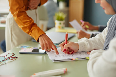 Caucasian woman standing and pointing at worksheet, while young adult Middle Eastern woman wearing...