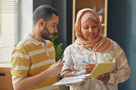Young adult Middle Eastern man and woman discussing notes and holding pens and notebooks in language school classroom, collaborating on educational activityの写真素材