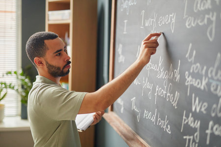 Young adult Black man writing English sentences on chalkboard in classroom holding notebook, demonstrating grammar concepts during language lesson at language schoolの写真素材