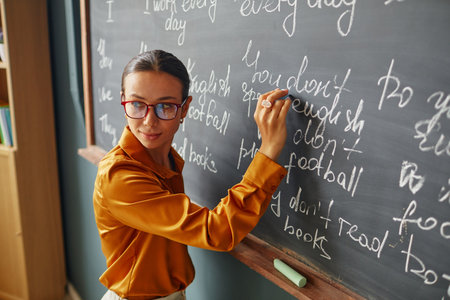 Caucasian young adult woman writing English phrases on chalkboard, wearing glasses, standing in...