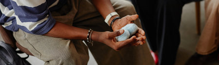 Header of Black man sitting holding blue stress ball in hands, wearing bracelet and watch, participating in group therapy session, partial view of another in background, focus on handsの写真素材