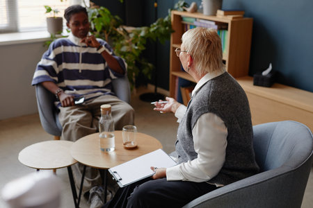 Middle aged Caucasian woman holding clipboard talking to young Black man sitting across in modern...