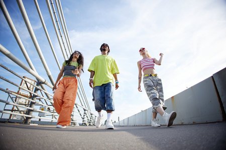 Group of three multiethnic people walking outdoors on urban bridge, smiling and talking enjoying...