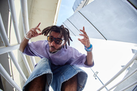 Portrait of male zoomer wearing sunglasses crouching outdoors, looking into camera with expressive...