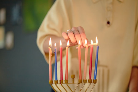 Woman lighting candles on menorah during Hanukkah celebration, hand carefully placing final candle...