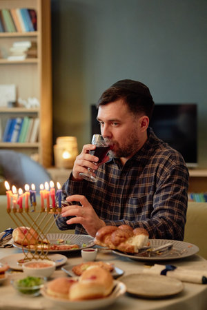 Caucasian young adult man wearing kippah sitting at table drinking wine during Hanukkah celebration...