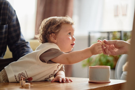 Caucasian child reaching out to take small object from adult hand during Hanukkah celebration, menorah visible in background, toddler showing curiosity and engagement at tableの写真素材