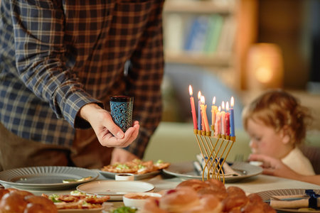 Man holding cup near menorah with lit candles on table while Caucasian child sitting in background eating during Hanukkah family celebration at homeの写真素材