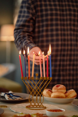 Man lighting candles on menorah during Hanukkah celebration, hand holding match near lit candles,...