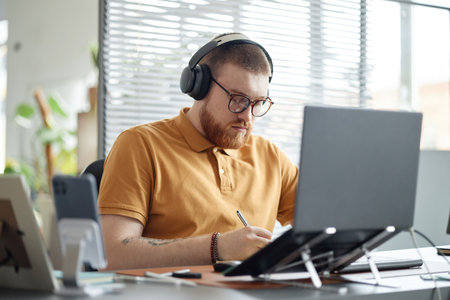 Young adult man wearing headphones working at desk using laptop and writing in notebook, focused...