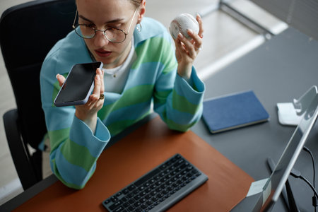 Caucasian young adult woman sitting at desk holding baseball in one hand and speaking into...