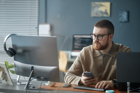 Young adult man with beard and glasses sitting at desk working on computer while holding smartphone,...