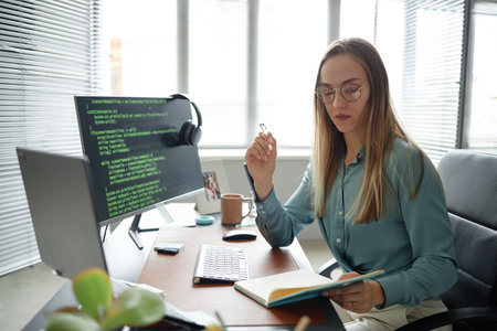 Young adult woman sitting at desk working on computer with code on screen, holding pen and notebook,...