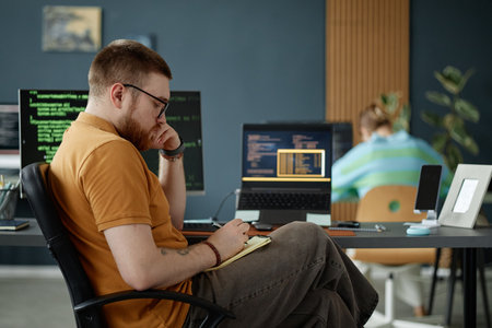 Young adult man with beard sitting at desk working on laptop with code on screen, holding pen and...