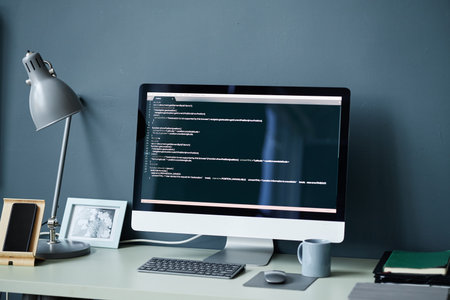 Modern desktop computer displaying programming code on screen standing on desk with table lamp,...