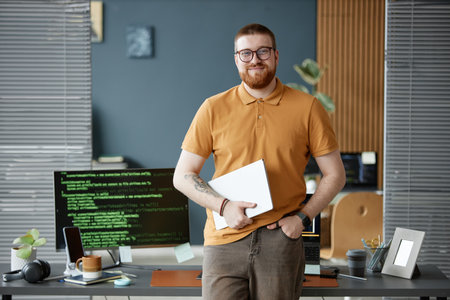 Caucasian young adult man with beard standing in modern office holding laptop, smiling confidently...