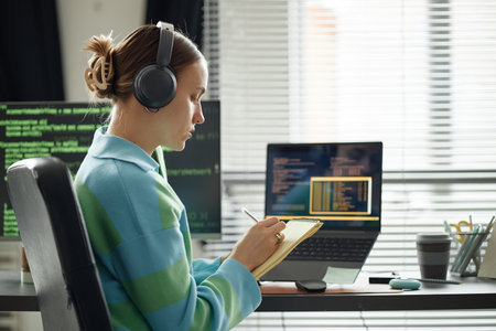 Caucasian young adult woman wearing headphones sitting at desk writing in notebook while working on...