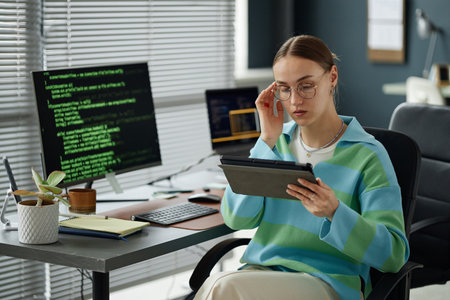 Young adult woman sitting at desk using digital tablet, adjusting eyeglasses with one hand, computer...